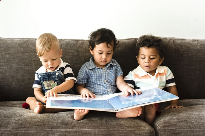 children, kids, boys, blonde, brunette, bare feet, toes, overalls, button-up, blue, polo shirt, couch, gray, brown, wall, white, book, picture book, reading